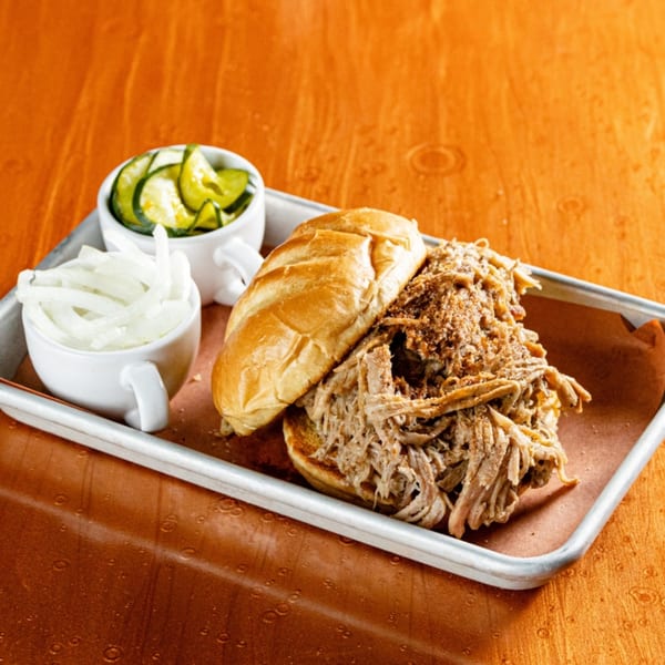 Reunion BBQ pulled pork sandwich with sliced onions and pickles on a metal tray.