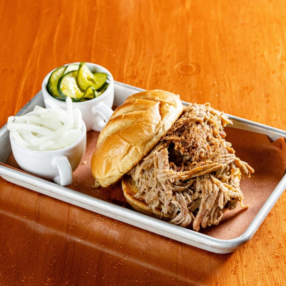 Reunion BBQ pulled pork sandwich with sliced onions and pickles on a metal tray.