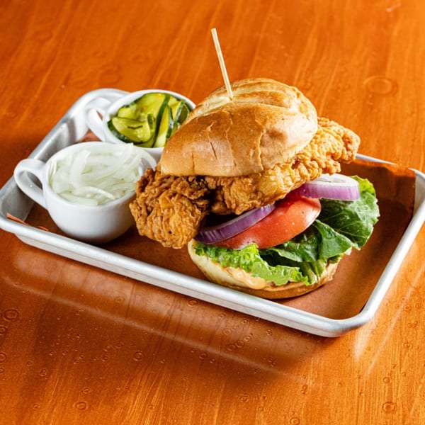 Crispy fried chicken sandwich with lettuce, tomato, red onion, and pickles on a metal tray.