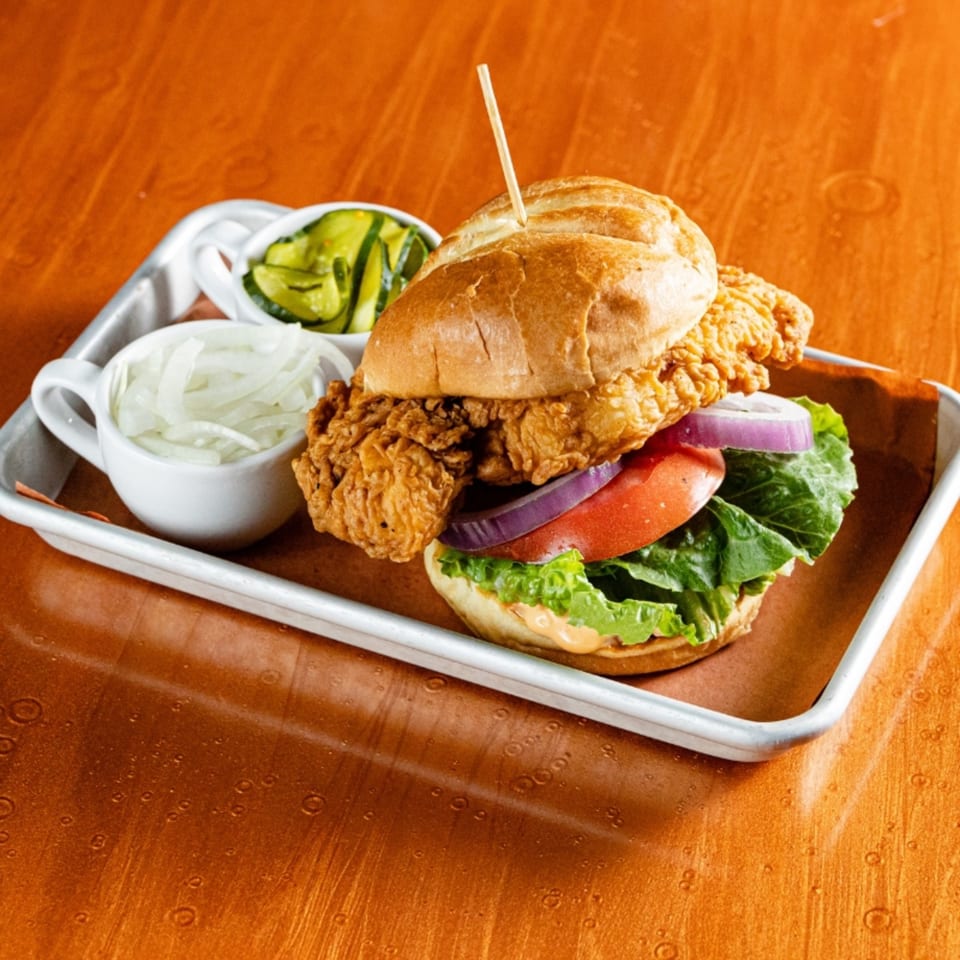 Crispy fried chicken sandwich with lettuce, tomato, red onion, and pickles on a metal tray.