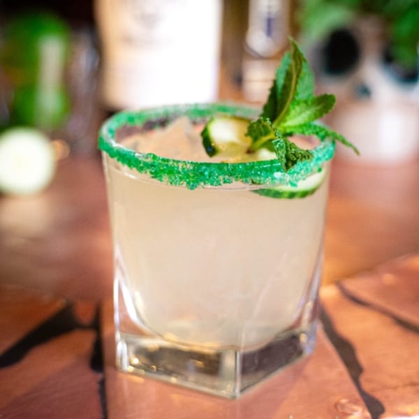 A cocktail with a green sugar rim, cucumber slices, and a mint sprig on a copper table.