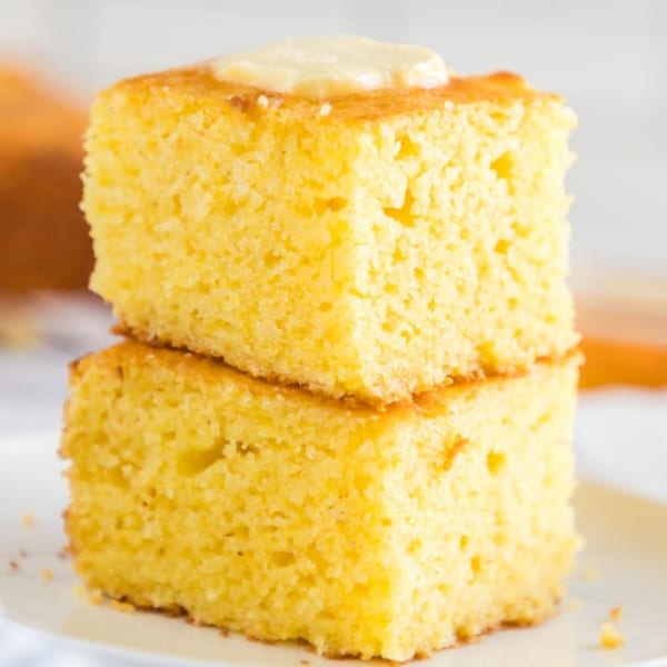 Two stacked squares of golden cornbread, the top piece topped with a dollop of butter.