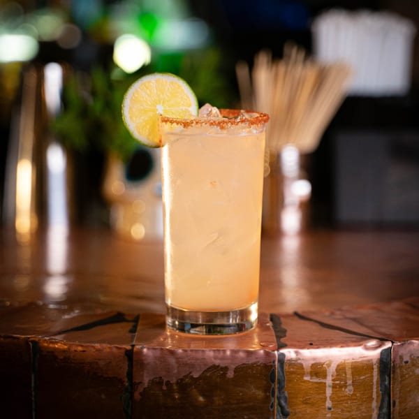 A tall, light-colored cocktail with a chili-salted rim and lime slice sits on a bar.