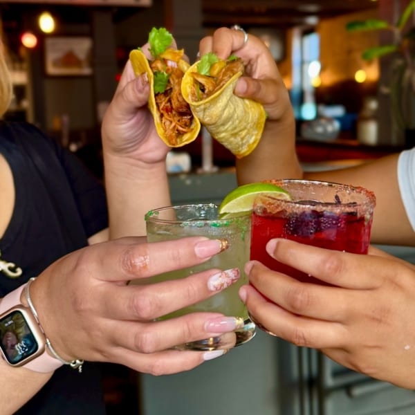 Two people clinking glasses of red and green drinks, holding Reunion BBQ tacos overhead.